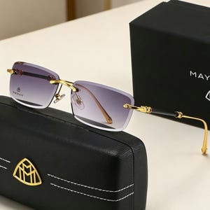 May include: Rimless sunglasses with gradient lenses, gold-tone metal frames, and black accents. The sunglasses rest on a black case with a gold emblem and are next to a black box with the word "MAY" visible.