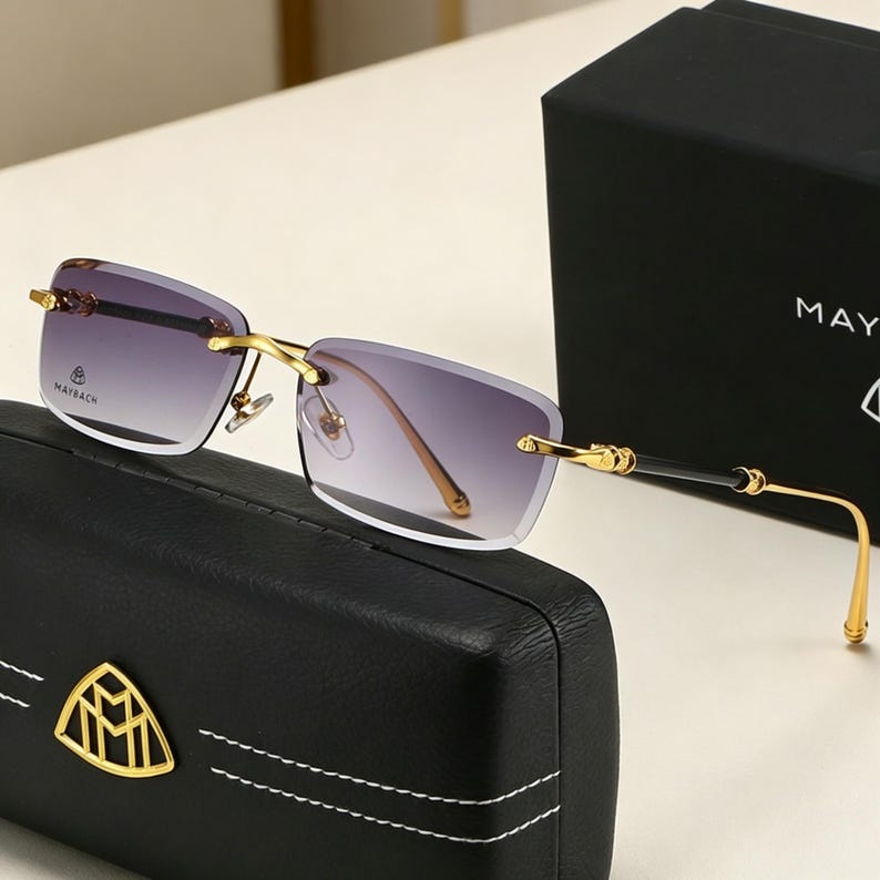 May include: Rimless sunglasses with gradient lenses, gold-tone metal frames, and black accents. The sunglasses rest on a black case with a gold emblem and are next to a black box with the word "MAY" visible.