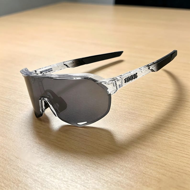 May include: Clear-framed sports sunglasses with dark gray lenses. The sunglasses have black arms and the brand name "100%" printed on the side. The lenses are a wraparound style, offering full coverage. The sunglasses are designed for athletic activities.
