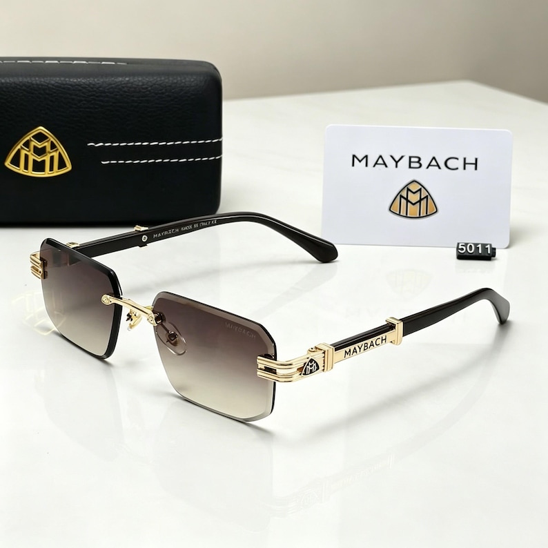 May include: Rimless sunglasses with gradient brown lenses and black temples. Gold-colored accents adorn the frame, with the brand name "MAYBACH" visible on the temples. A black case and a white card with the brand logo are also present.