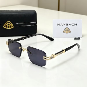 May include: Black rimless sunglasses with gold accents and dark lenses. The temples are black with gold detailing and the brand name "MAYBACH". A black case and a white card with the brand logo and name are also visible.