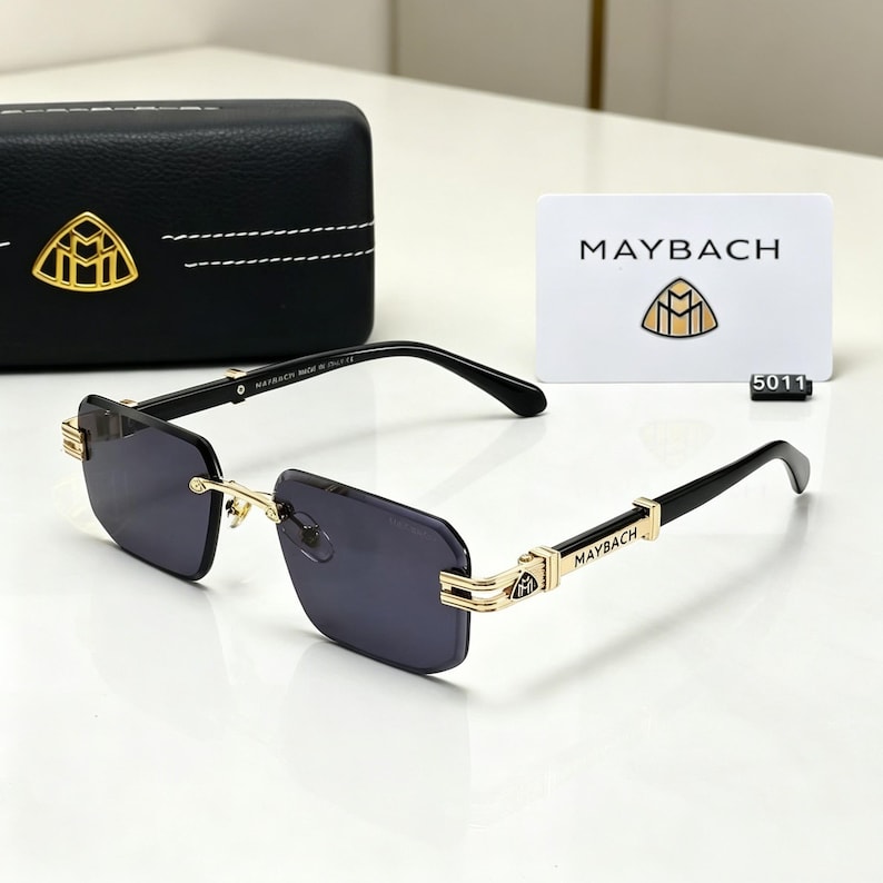 May include: Black rimless sunglasses with gold accents and dark lenses. The temples are black with gold detailing and the brand name "MAYBACH". A black case and a white card with the brand logo and name are also visible.