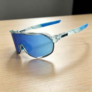 May include: Blue-tinted sports sunglasses with a clear frame. The lenses are a vibrant blue, and the arms transition from clear to blue. The text "100%" is printed on the arm. The sunglasses are designed for sports and outdoor activities.
