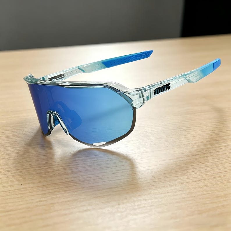 May include: Blue-tinted sports sunglasses with a clear frame. The lenses are a vibrant blue, and the arms transition from clear to blue. The text "100%" is printed on the arm. The sunglasses are designed for sports and outdoor activities.