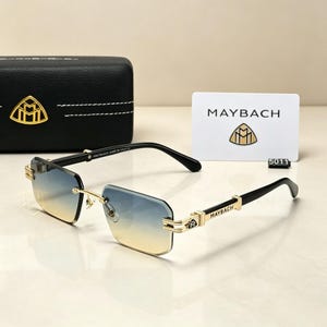 May include: Rimless sunglasses with gradient blue and yellow lenses, gold-tone accents, and black temples. The brand name "MAYBACH" is visible on the temples and a white card. A black case with a gold logo is also present.