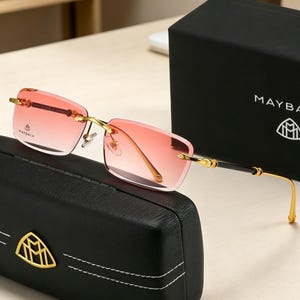 May include: Rimless sunglasses with gold-tone metal frames and gradient pink lenses. The sunglasses rest on a black leather case with a gold emblem. A black box with the brand name "MAYBACH" is in the background.