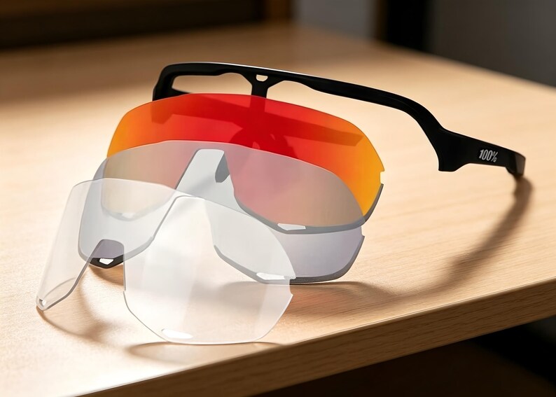 May include: A set of interchangeable sports sunglasses with a black frame and multiple lenses. The lenses include a vibrant red-orange, a mirrored silver, and clear options. The frame has the text "100%" printed on the arm.