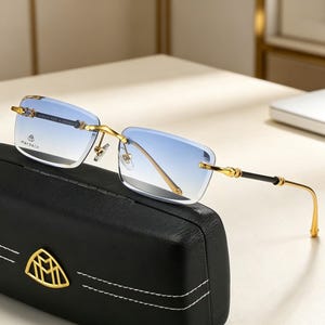 May include: Rimless sunglasses with gradient blue lenses and gold-tone metal frames. The temples have black accents and decorative details. The glasses rest on a black leather case with a gold emblem. The brand name "MAYBACH" is visible on the lens.
