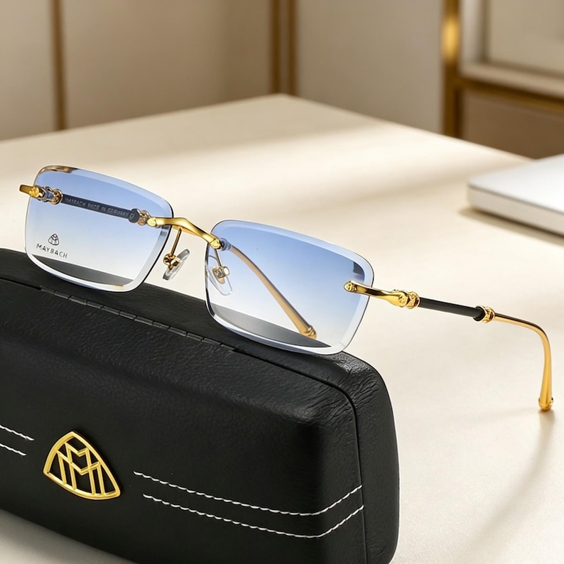 May include: Rimless sunglasses with gradient blue lenses and gold-tone metal frames. The temples have black accents and decorative details. The glasses rest on a black leather case with a gold emblem. The brand name "MAYBACH" is visible on the lens.