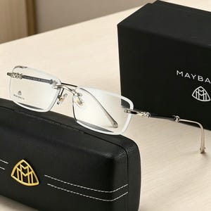 May include: Rimless eyeglasses with silver frames and black temples, resting on a black leather case with a gold emblem. The glasses are next to a black box with the brand name "MAYBACH" and logo.