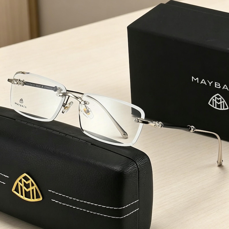 May include: Rimless eyeglasses with silver frames and black temples, resting on a black leather case with a gold emblem. The glasses are next to a black box with the brand name "MAYBACH" and logo.