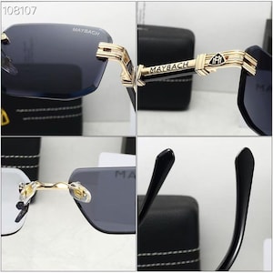 May include: Rimless sunglasses with dark gray lenses and gold-colored accents. The temples feature the brand name "MAYBACH" and a logo. The sunglasses have a sleek, modern design.