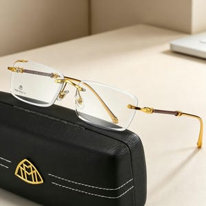 May include: Rimless eyeglasses with gold-colored frames and temples, resting on a black leather case. The glasses have a rectangular lens shape and the brand name "MAYBACH" is visible on the lens.