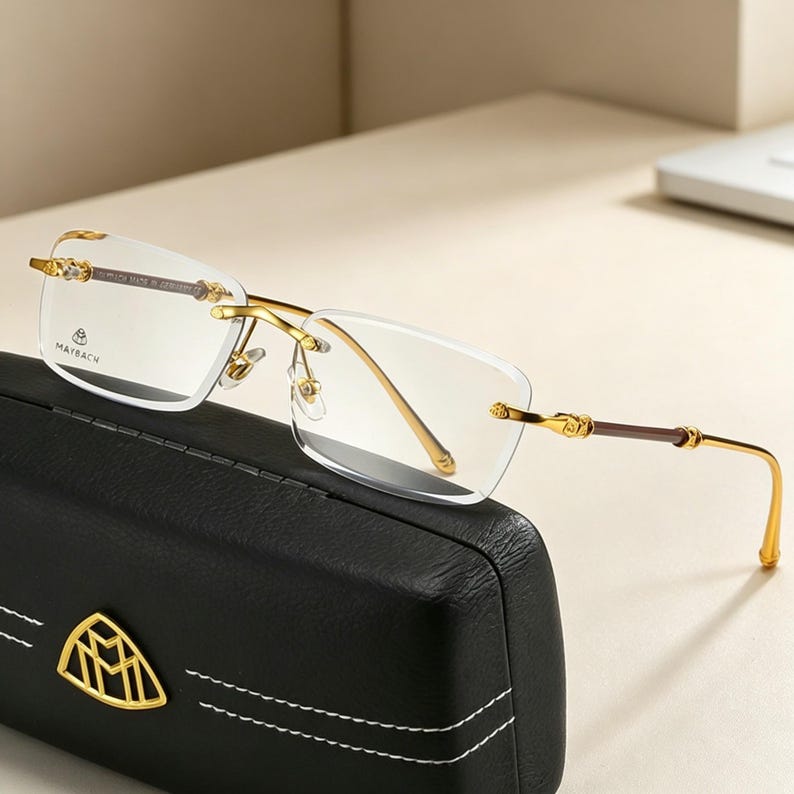 May include: Rimless eyeglasses with gold-colored frames and temples, resting on a black leather case. The glasses have a rectangular lens shape and the brand name "MAYBACH" is visible on the lens.