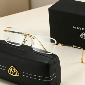May include: Rimless eyeglasses with gold-tone metal frames and temples, resting on a black leather case. The glasses have clear lenses and the brand name "MAYBACH" is visible on the lens and case. A black box with the brand name is in the background.