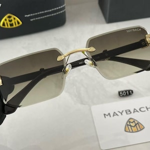 May include: Rimless sunglasses with gradient lenses. The sunglasses have a gold-colored bridge and accents, with the brand name "MAYBACH" visible on the top of the lenses. A small tag with the number "5011" is also present.
