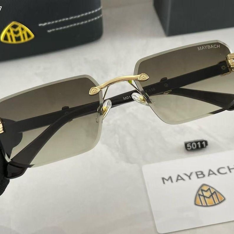 May include: Rimless sunglasses with gradient lenses. The sunglasses have a gold-colored bridge and accents, with the brand name "MAYBACH" visible on the top of the lenses. A small tag with the number "5011" is also present.