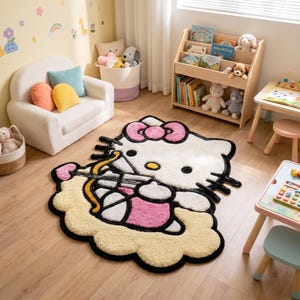 Może przedstawiać: Pluszowy dywanik w kształcie postaci Hello Kitty, z białą twarzą, dużą różową kokardą, żółtym nosem i czarnymi wąsami. Postać trzyma żółty łuk ze strzałą i spoczywa na miękkiej podstawie w kształcie jasnej, żółtej chmury.
