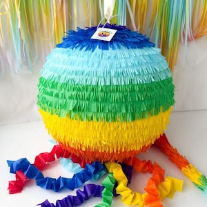 Może przedstawiać: Kolorowa, okrągła piñata z warstwami niebieskich, zielonych, żółtych i pomarańczowych frędzli. Tęczowe serpentyny i pasujący ogon wychodzą z dołu. Biała wstążka i etykieta są przymocowane na górze.