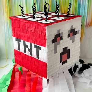 Puede incluir: Una piñata cuadrada con un diseño de bloque TNT rojo y blanco, letras negras en un lado y una cara pixelada blanca en el otro. La parte superior tiene detalles de limpiapipas negros y cuelgan largas serpentinas de papel rojo, verde y blanco.