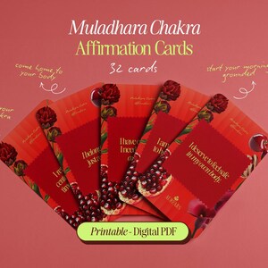 Könnte beinhalten: Ein Set aus 32 Muladhara Chakra Affirmationskarten in Fächerform. Die Karten haben rote Hintergründe, Granatapfel-Illustrationen und goldenen Text. Handschriftliche Notizen enthalten Phrasen wie 'balance your root chakra' und 'heal your root'.