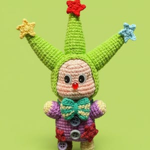 May include: A handmade crochet doll in a whimsical jester design. The doll has a green pointed hat with star accents, a pink face, and a purple and red outfit. It has a green bow tie and button details.