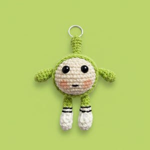 May include: A handmade crochet keychain in the shape of a green and cream-colored creature with black eyes and rosy cheeks. The keychain has a small loop at the top and features small arms and legs with black stripes. The background is a solid light green.