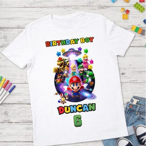 May include: White t-shirt with a vibrant Super Mario Bros. graphic. The design includes Mario and other characters, with the text "BIRTHDAY BOY", the name "DUNCAN", and the number "6".