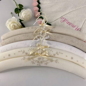 Peut inclure: Un ensemble de trois cintres rembourrés en tissu beige et blanc, ornés de broderies florales délicates, de petits nœuds en ruban de satin au niveau du crochet et de crochets en métal argenté. Les cintres sont disposés verticalement avec deux roses artificielles blanches.
