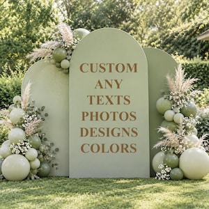 Può includere: Uno sfondo ad arco color verde salvia con il testo marrone CUSTOM ANY TEXTS PHOTOS DESIGNS COLORS. L'espositore è decorato con grappoli di palloncini verde salvia di varie dimensioni, erba della pampa essiccata e piccoli fiori bianchi.