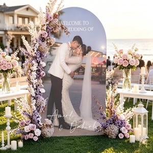 May include: An arched wedding welcome sign featuring a photo of a couple, with the text "WELCOME TO THE WEDDING OF" and "Amerl & Dahirb" in white script. The sign is decorated with large floral arrangements of purple and pink roses.