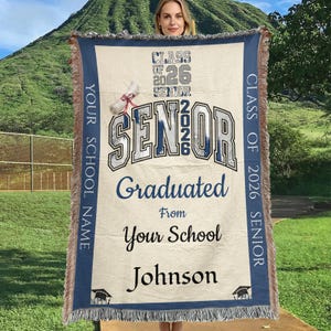 Puede incluir: Una manta tejida beige con ribete y flecos azules. La manta presenta el texto "CLASS OF 2026 SENIOR" y "SENIOR Graduated From Your School Johnson" en azul y blanco. Un gráfico de gorro de graduación está en la parte inferior.