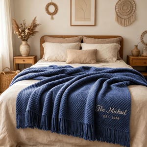 Puede incluir: Una manta azul marino texturizada con flecos, que presenta un bordado blanco con el texto The Michael EST. 2026. La manta está colocada sobre una cama con ropa de cama beige, mostrando un patrón repetitivo de diamantes en relieve en la tela.
