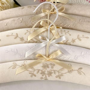 May include: A set of padded clothes hangers covered in beige and white fabric, featuring delicate floral embroidery in matching thread tones. Each hanger includes a metal hook wrapped in coordinating ribbon and is finished with a small satin bow.