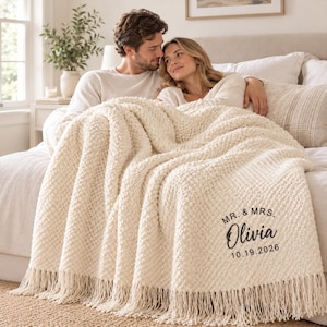 May include: A cream-coloured, chunky knit throw blanket featuring a textured popcorn stitch pattern and long fringe along the bottom edge. The blanket is personalised with black embroidered text that reads MR. & MRS. Olivia 10.19.2026.