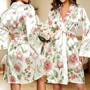 May include: A white satin robe featuring a pink floral pattern with green leaves and the name Annie printed on the back. The robe has long sleeves, a wrap-around front with a matching tie belt, and a shawl collar. The fabric has a smooth, glossy finish.