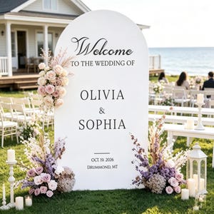 Può includere: Un cartello di benvenuto per matrimoni bianco a forma di arco con testo nero che recita Welcome to the wedding of Olivia & Sophia, Oct. 19, 2026, Drummond, MT. Il cartello è decorato con composizioni di rose rosa, fiori viola ed erbe essiccate.