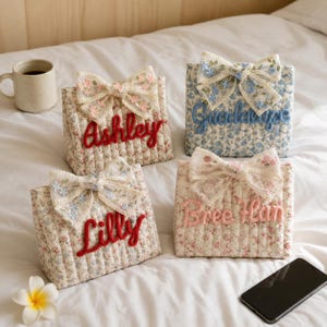 Könnte beinhalten: Vier gesteppte Stofftaschen mit Blumenmustern, jede mit einer dekorativen Spitzenschleife und einem in Schreibschrift gestickten Namen versehen: Ashley, Guadalupe, Lilly und Bree Han. Die Taschen sind rechteckig und haben vertikale Nähte.