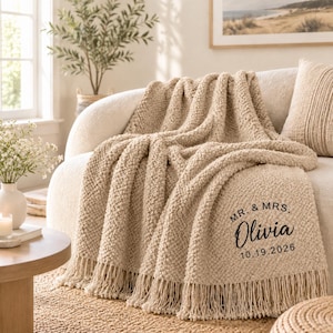 Könnte beinhalten: Eine beige, strukturiert gestrickte Wohndecke mit Fransenbesatz, die in der unteren rechten Ecke den schwarzen Aufdruck "MR. & MRS. Olivia 10.19.2026" trägt. Die Decke hat ein dickes, genopptes Webmuster und liegt über einem geschwungenen Sofa.