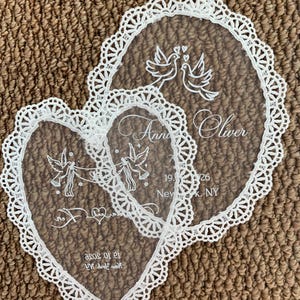 May include: Two white lace-trimmed fabric pieces, one oval and one heart-shaped. The oval piece features two doves and the text "Anne Oliver 19.10.2026 New York, NY". The heart-shaped piece shows two doves holding a banner with text and dates.