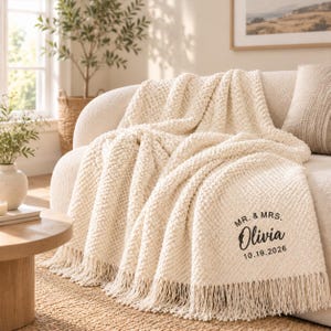 May include: A cream-coloured knitted throw blanket with a textured popcorn stitch pattern and fringe trim. The blanket features black printed text that reads MR. & MRS. Olivia 10.19.2026, positioned on the lower right corner of the fabric.