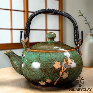 Op de afbeelding: Een keramische theepot met een groen en bruin glazuur, met een zwart geweven handvat en bloemmotieven. De theepot heeft een tuit en een deksel met een kleine knop. De merknaam "HIKARYCLAY" staat rechtsonder.