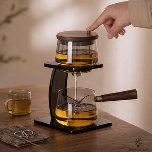 May include: A glass tea brewing set featuring a dark wood lid and handle, mounted on a black stand. The upper glass vessel holds amber tea, which flows through a central spout into a lower glass pitcher. A small glass cup sits nearby.