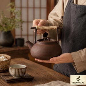 May include: A brown ceramic teapot with a rope handle is held in the hands. The teapot has a round body with horizontal ridges. A small teacup with vertical stripes sits on a wooden tray. The image also includes the text "光 HIKARYCLAY".