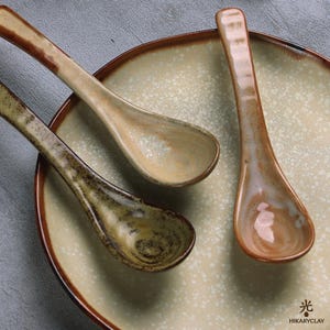 May include: Two ceramic spoons with a speckled beige and brown glaze rest on a matching round plate. The spoons feature a curved handle and a deep bowl. The plate has a dark brown rim and a textured, light-colored center. Text reads HIKARYCLAY.