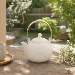 May include: A cream-coloured ceramic teapot with a wooden handle and lid. The teapot has a rounded shape with a wavy design. The handle is made of light-coloured wood. The teapot is on a wooden table with a cup and saucer.