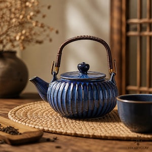 May include: A blue ceramic teapot with a ribbed design, a dark brown woven handle, and a matching lid. The teapot is on a woven mat, with a teacup visible to the right. The background includes a wooden table and a vase with dried flowers.
