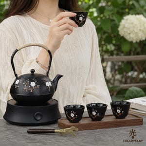 Può includere: Una teiera in ghisa nera con superficie testurizzata e manico rivestito, decorata con motivi di fiori di prugno bianchi. Il set include tre tazze da tè nere abbinate su un vassoio di legno e un utensile in metallo con manico.