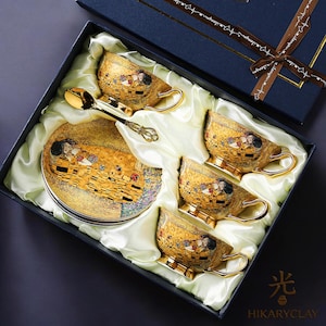 May include: A boxed set of gold-colored teacups, saucers, and spoons. The teacups and saucers feature a famous artwork design. The set includes four teacups, four saucers, and four spoons, all presented in a navy blue gift box.