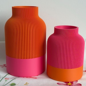 Könnte beinhalten: Zwei dekorative Vasen. Die größere Vase ist orange mit einem rosa Sockel, und die kleinere Vase ist rosa mit einem orangenen Sockel. Beide Vasen haben eine gerippte Textur und eine abgerundete Form. Die Vasen stehen auf einer Oberfläche mit floralem Muster.